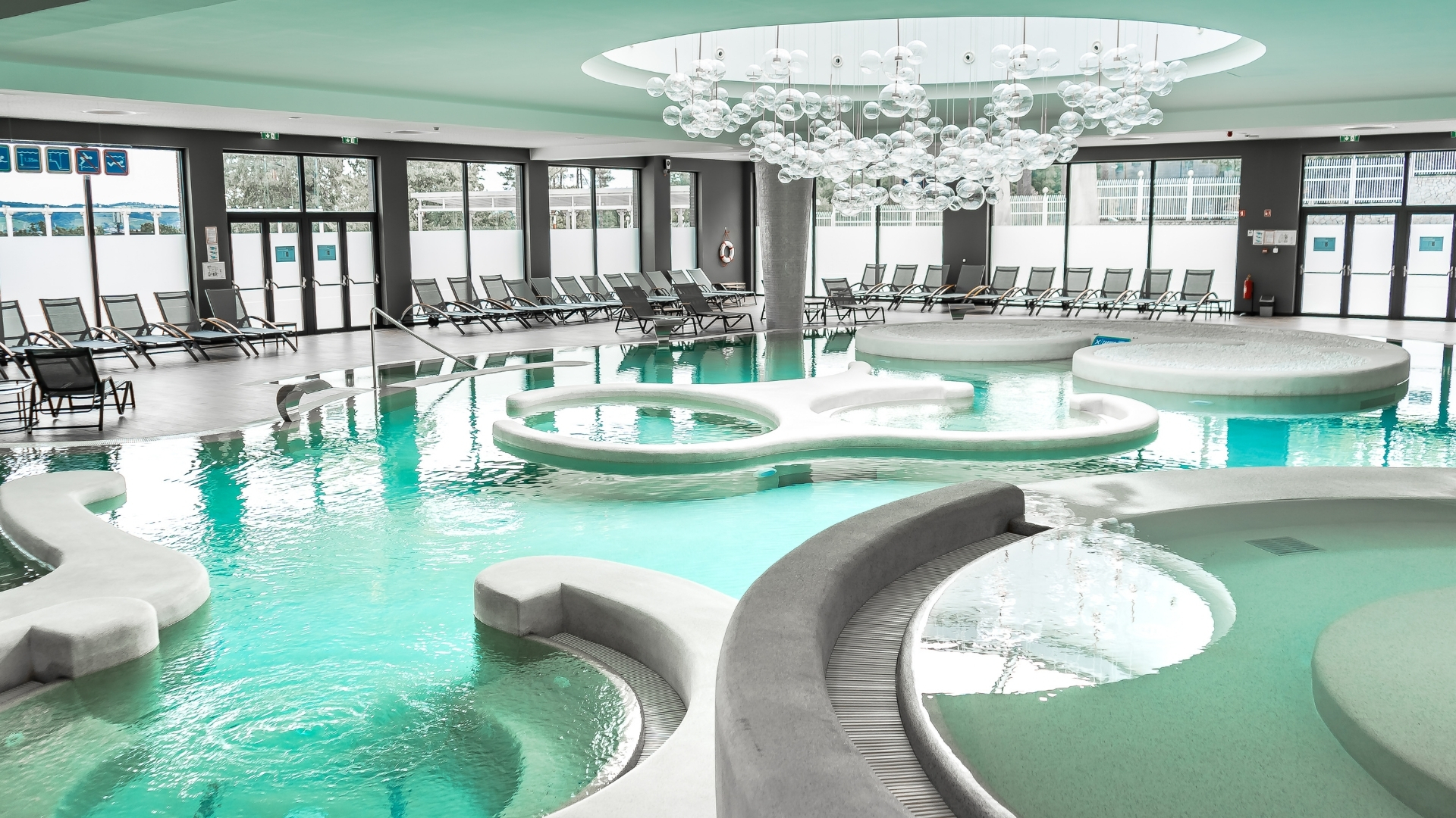 Ein weitläufiger Thermal-Innenbereich mit verschiedenen organisch geformten Becken und einer markanten Skulptur aus Glaskugeln an der Decke.