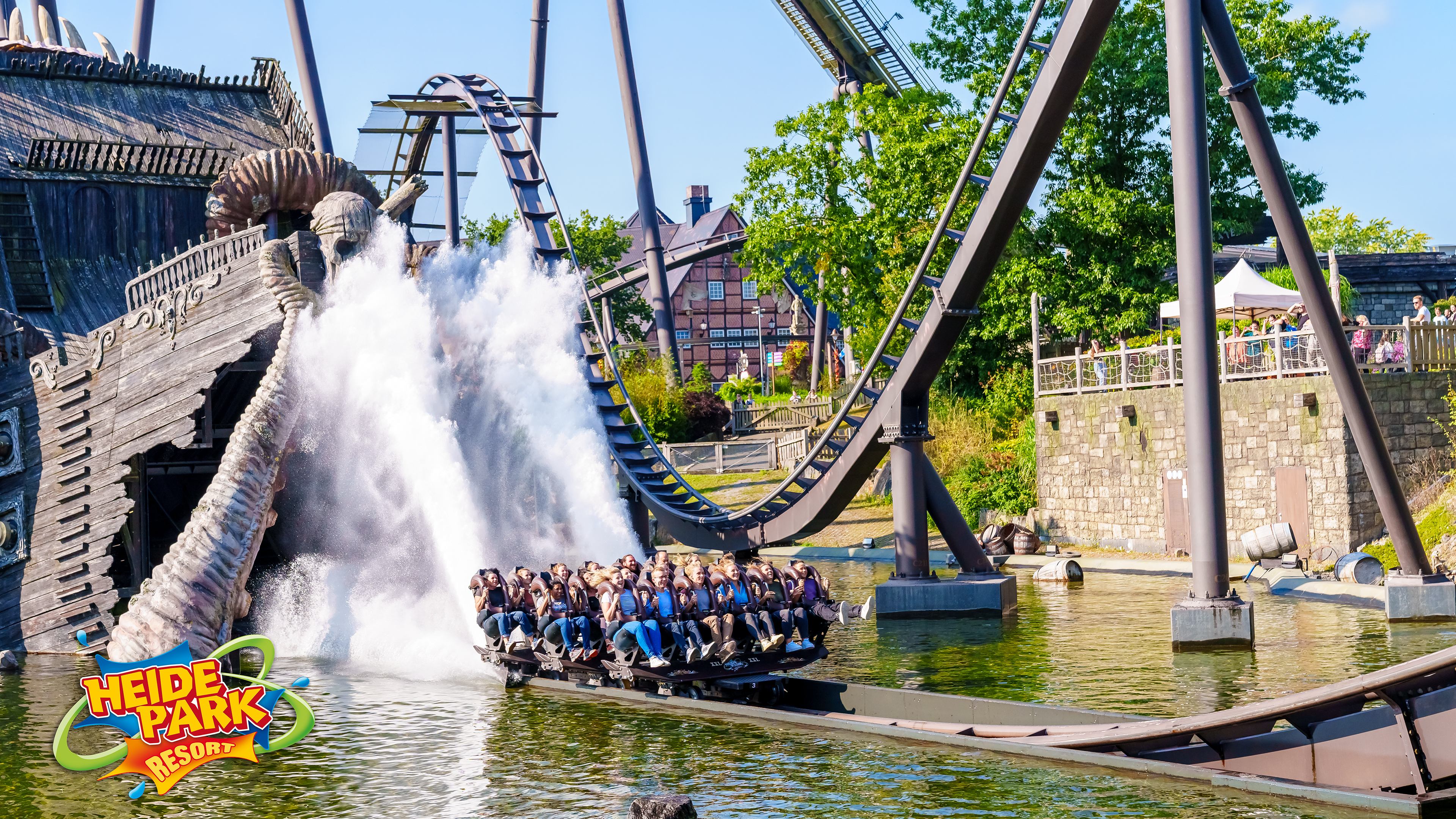 Ein großes Boot einer Wasserattraktion im Heide Park Resort rutscht eine Schiene hinunter und verursacht beim Eintauchen in den See eine gewaltige Wasserfontäne. Fahrgäste sitzen jubelnd im Boot. Im Hintergrund sind die Schienen einer großen dunklen Achterbahn und ein Fachwerkhaus zu erkennen.