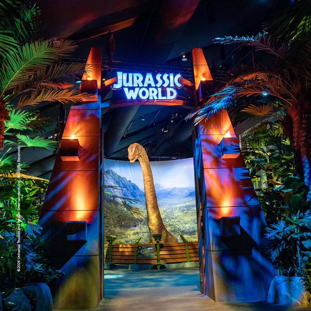 Blick auf das ikonische Eingangstor der „Jurassic World Exhibition“. Durch das Tor sieht man den langen Hals eines Brachiosaurus vor einer Kulisse aus Bergen und Urwald. Die Szene ist von Palmen und tropischen Pflanzen umgeben.