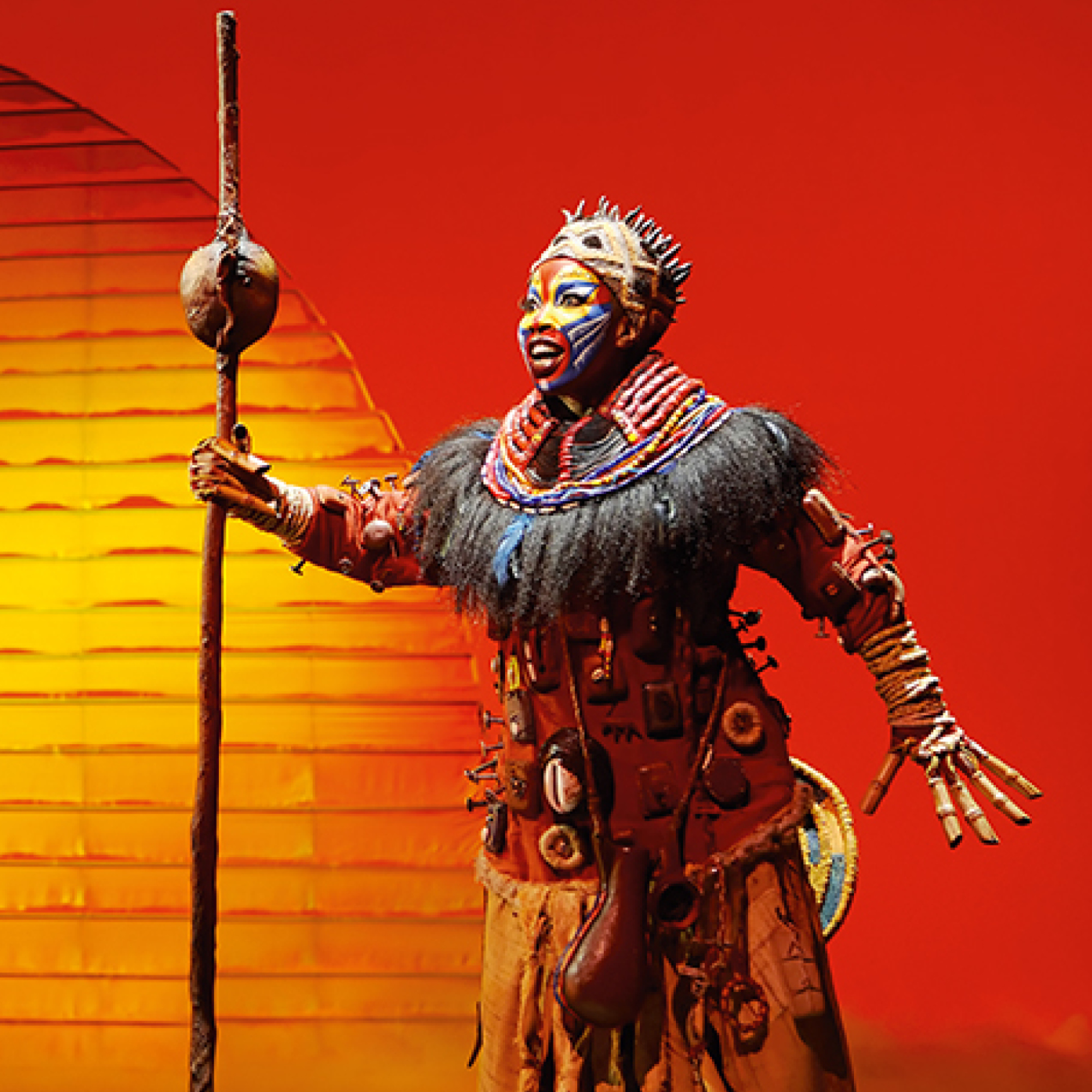 Eine beeindruckende Aufnahme der Figur Rafiki aus dem Musical „Der König der Löwen“. Die Darstellerin trägt ein detailreiches Kostüm mit traditionellem Kopfschmuck und kunstvoller Gesichtsbemalung. Sie hält einen langen Holzstab in der Hand und singt mit leidenschaftlichem Ausdruck. Der Hintergrund wird von einer riesigen, strahlend gelben Sonne vor einem tiefroten Abendhimmel dominiert, was die Atmosphäre der afrikanischen Savanne heraufbeschwört.