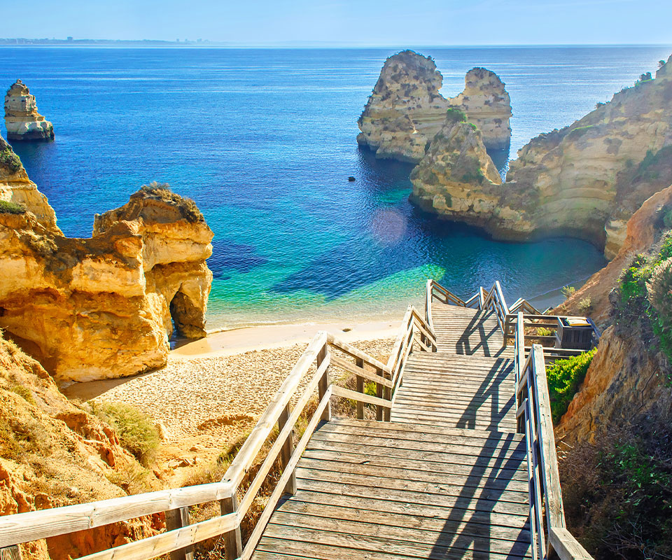 Urlaub Portugal. Holzsteg der runter zum Strandabschnitt führt. Hellbraune Felsen drumherum und im Meer.